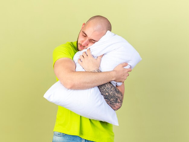 Sleepy young ill man hugs and puts head on pillow isolated on olive green wall