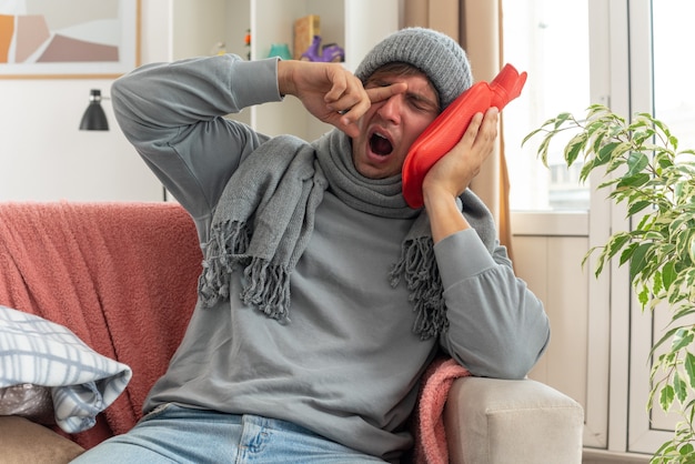 Sleepy young ill man with scarf around neck wearing winter hat wipes his eye with finger and holding hot water bottle sitting on couch at living room