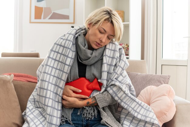 Free photo sleepy young ill slavic woman with scarf around her neck wrapped in plaid holding hot water bottle sitting on couch at living room