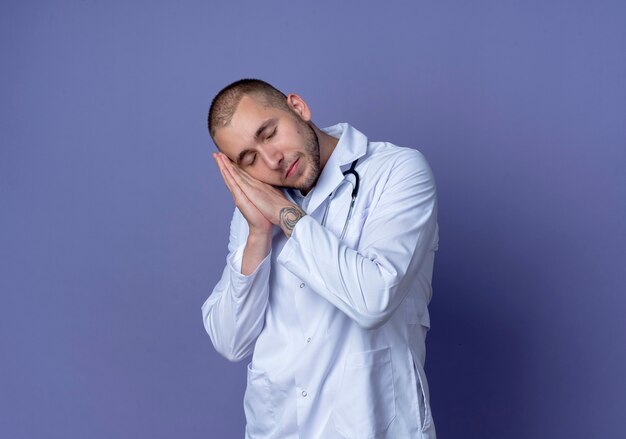 Sleepy young male doctor wearing medical robe and stethoscope doing sleep gesture with closed eyes isolated on purple wall