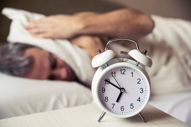 Sleepy young man covering ears with pillow as he looks at alarm clock in bed. sleeping man disturbed by alarm clock early morning. Frustrated man listening to his alarm clock while relaxing on his bed