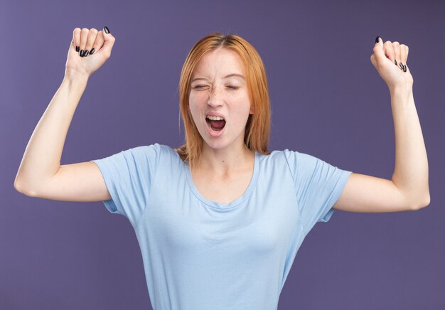 Sleepy young redhead ginger girl with freckles standing with raised fists and yawning isolated on purple wall with copy space