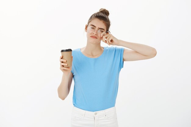 Сонная молодая женщина в очках пьет кофе и устала протирать глаза