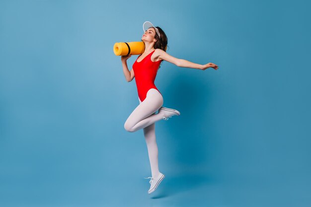 Slender dark-haired lady in red leotard and white pantyhose is holding yoga mat