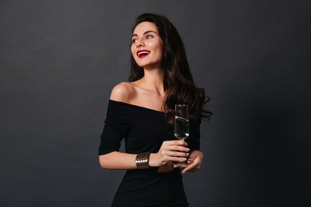 Стройная темноволосая дама в маленьком черном платье и стильном золотом браслете улыбается, держа бокал вина на изолированном фоне.