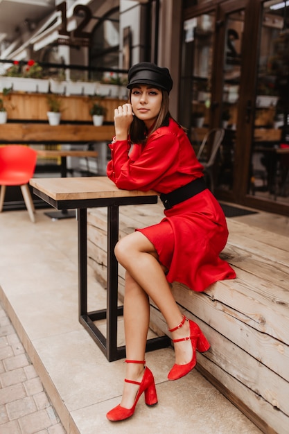 Slender girl in elegant shoes and belted dress sitting in cafe. Portrait of young woman looking thoughtfully