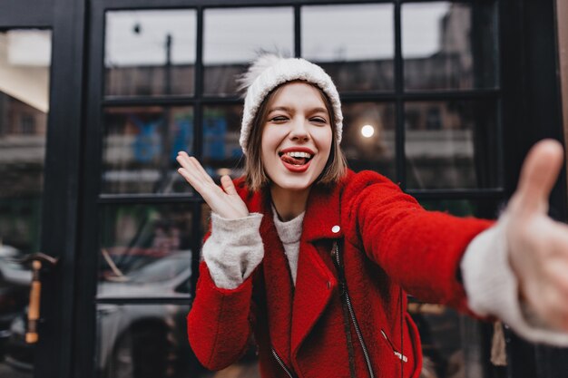 Slender girl in red coat shows her tongue, winks and takes selfie on background of window with black frame.