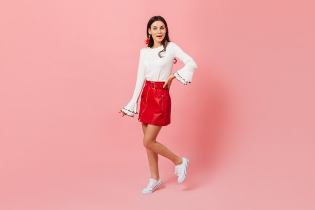 Slender girl in stylish white-red outfit posing on isolated background. Shot of brunette in great mood.