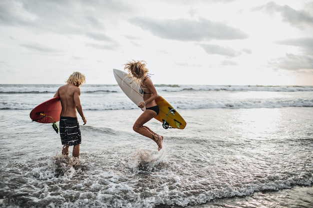 Бесплатное фото Стройный парень и девушка прыгают в морской воде и держат доски для серфинга