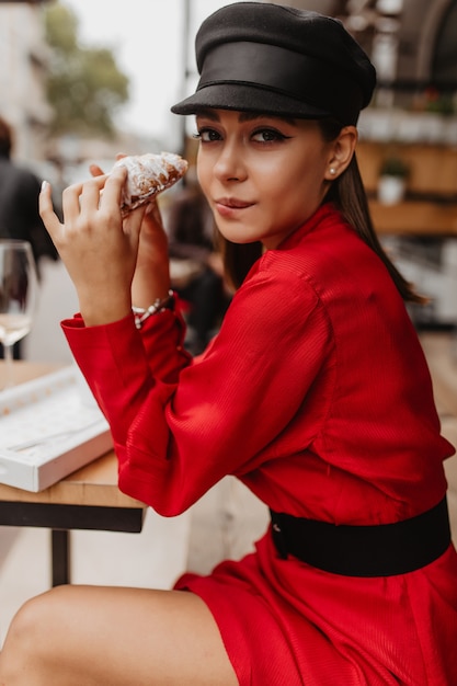 Стройная модель не сдержалась перед вкусным сладким круассаном, сидя в парижском кафе. Портрет молодой леди в красном на открытом воздухе