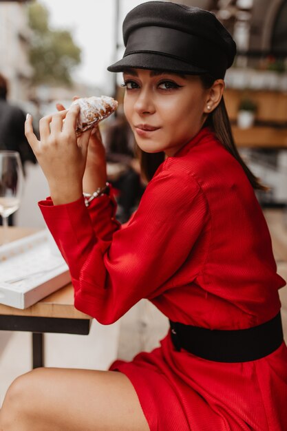 Стройная модель не сдержалась перед вкусным сладким круассаном, сидя в парижском кафе. Портрет молодой леди в красном на открытом воздухе