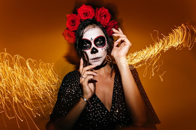 Free photo slender stylish woman with roses in her hair and mask of skull posing for festive portrait moving her hands gracefully