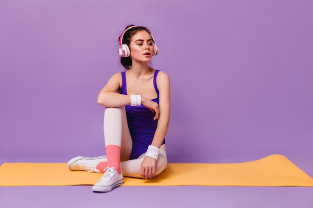 Slender woman in purple sports bodysuit and white headphones resting sitting on mat for fitness