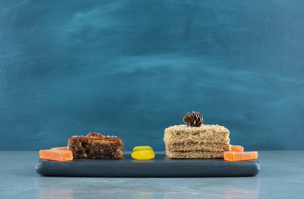 A slice of cake, bakhlava and some jelly candies on a navy board on marble surface