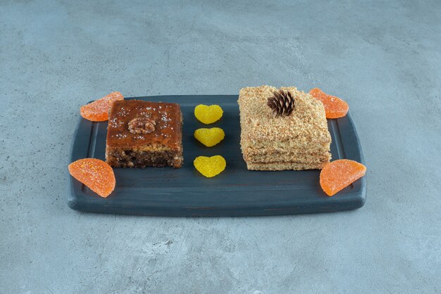 A slice of cake, bakhlava and some jelly candies on a navy board on marble surface