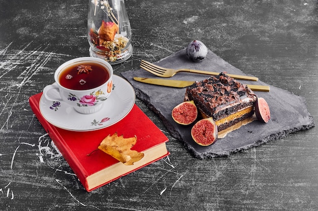 A slice of chocolate cake with fruits and a cup of tea on a stone platter. 