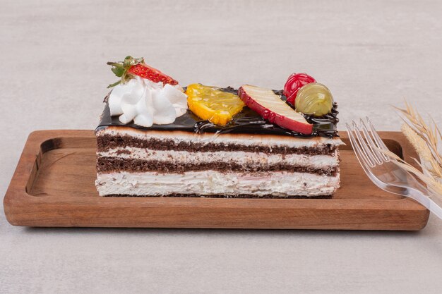 Slice of chocolate cake on wooden board with fruit slices.