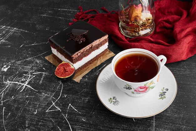 A slice of chocolate cheesecake on a black surface with figs and a cup of tea. 