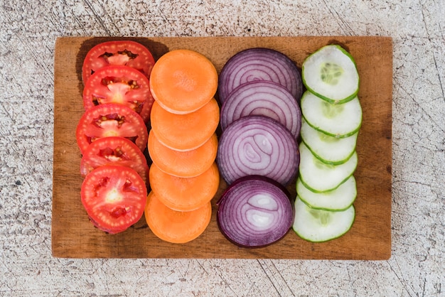 Slice of tomatoes; carrots; onion and cucumber arranged on wooden board over the concrete backdrop