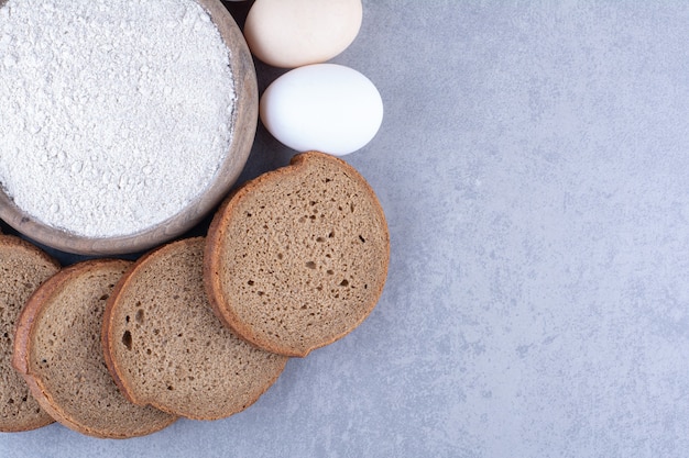 Sliced black bread and eggs around a bowl of flour on marble surface