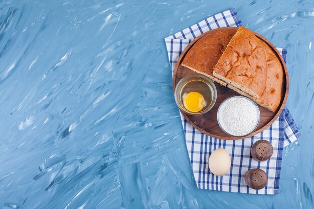 Sliced bread on a board next to flour and egg on a tea towel , on the blue.