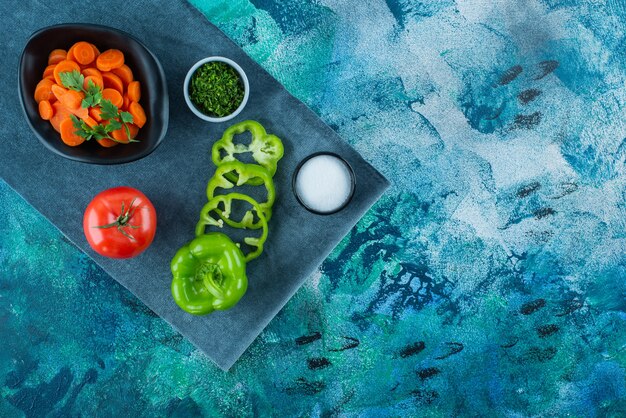 Sliced carrots in a bowl next to vegetables on a towel , on the blue background. 