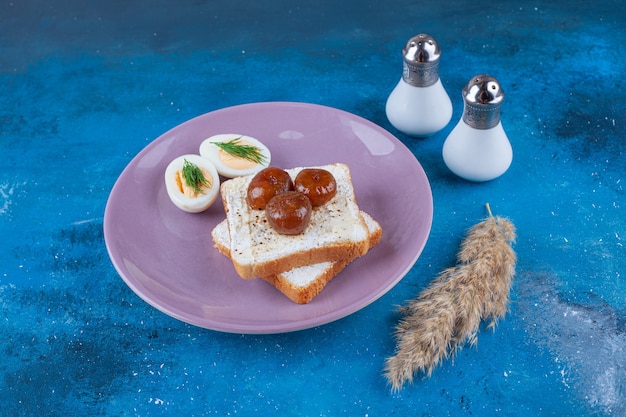 Нарезанное яйцо и варенье на сырном хлебе на тарелке, на синей поверхности.