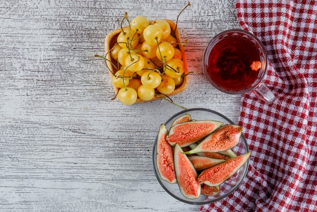 Sliced figs in a bowl with cherries, cup of tea top view on grungy and kitchen towel