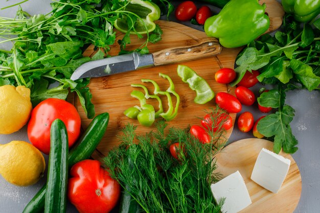 Sliced green pepper with tomatoes, salt, cheese, lemon, greens, knife on a cutting board on gray surface