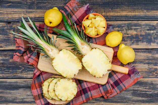 Sliced juicy pineapples with two quinces and a lemon in a wood boards and bowl on old wood grunge surface and picnic cloth, flat lay.
