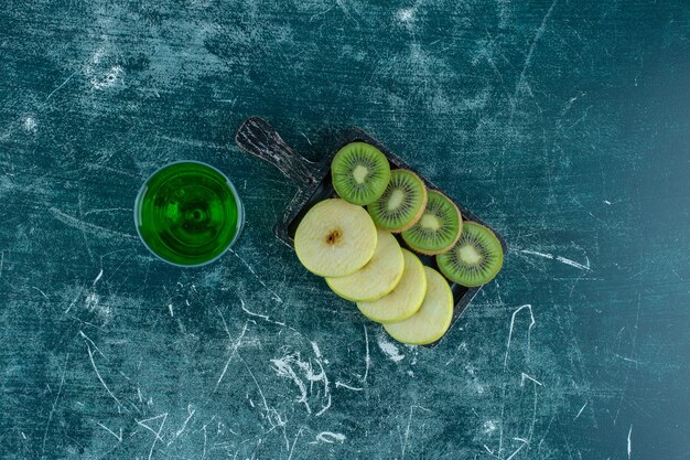 Sliced kiwi and apple on the board next to tarragon juice , on the blue table. 