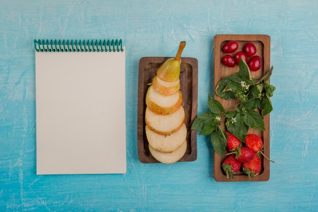 Sliced pears with strawberries and mulberries in wooden platters with a notebook aside