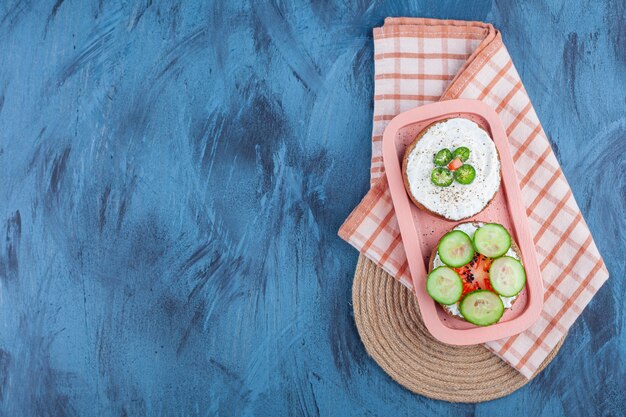 Sliced vegetables on cheese breads in board on towel on trivet on blue. 