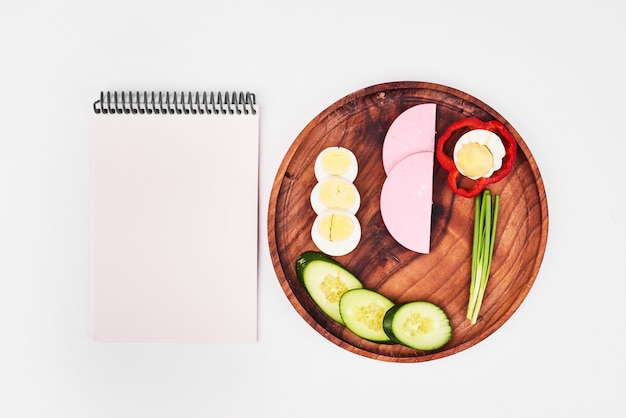 Slices of boiled egg, sausage, green onion, red pepper and cucumbers on wooden plate with notebook aside.