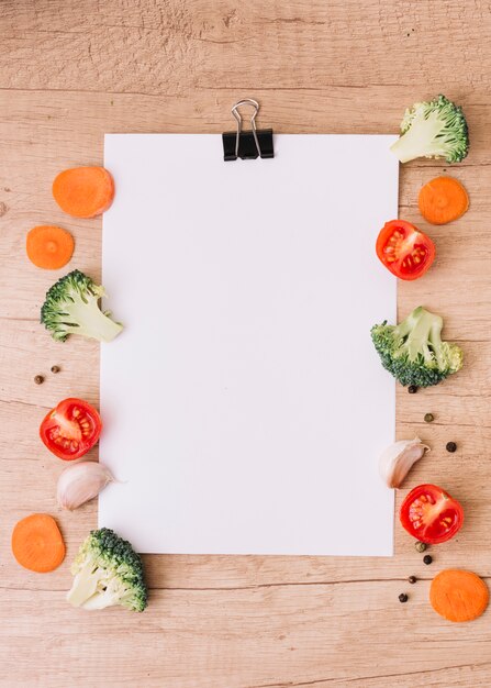 Slices of carrot; halved broccoli; tomatoes; garlic clove and black pepper on the side of blank white paper over the wooden desk