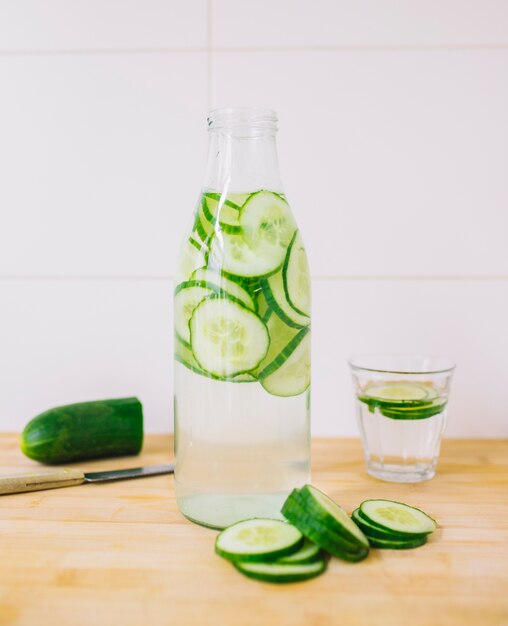 Slices of cucumber in the bottle of water and glass on wooden desk against wall