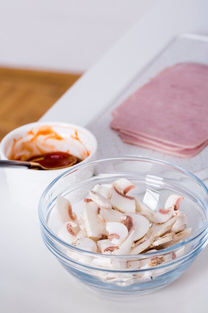 Slices of mushroom in the glass bowl; ham and sauce on table