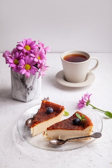 ブルーベリー​と​ミント​の​葉​と​プレート上​の​自家製​バスク焼き​チーズ​ケーキ​の​スライス​、​花瓶​の​花​と​明るい​背景​の​紅茶​の​カップ