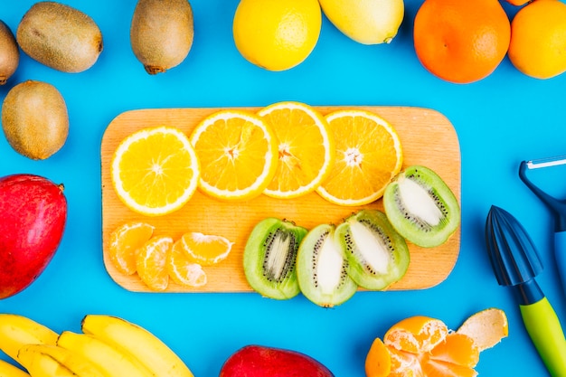 Slices of oranges and kiwi on chopping board surrounded with fruits on blue background