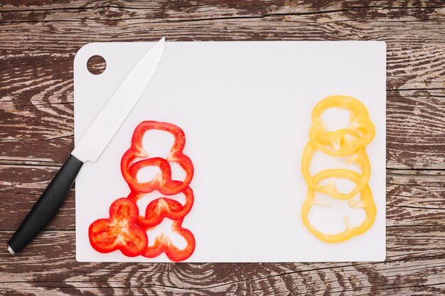 Slices of red and yellow bell pepper on white chopping board over the wooden texture background