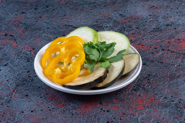 Slices of zucchini, eggplant and bell pepper with a small parsley bundle on a platter on dark colored background. High quality photo