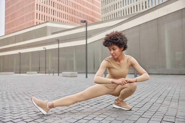 Foto gratuita la donna snella e attiva in abbigliamento sportivo fa esercizi di squat allunga le gambe indossa scarpe da ginnastica si riscalda prima che l'allenamento si metta all'aperto contro gli edifici della città alta. persone sport e stile di vita sano concetto