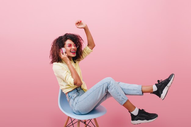 Slim african woman with long legs listening music and laughing. Stunning mulatto female model in black shoes sitting on blue chair.