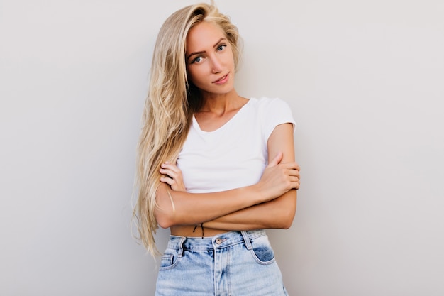 Slim attractive woman in white tank-top expressing interest. glad blonde woman standing on light background.