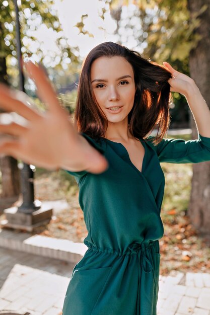 Slim attractive young woman with dark hair wearing green dress and touching her hair and waving at camera by hand