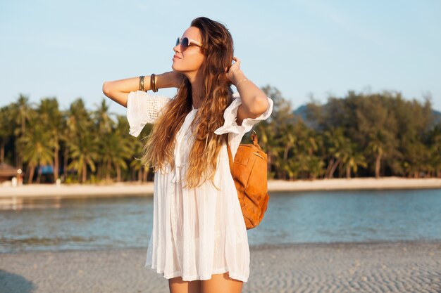 Slim beautiful woman in white cotton dress walking on tropical beach on sunset holding leather backpack.