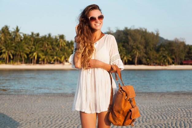 Худенькая красивая женщина в белом платье на тропическом пляже на закате, держа кожаный рюкзак.
