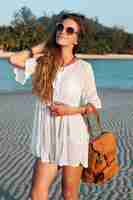 Free photo slim beautiful woman in white dress on tropical beach on sunset holding leather backpack.