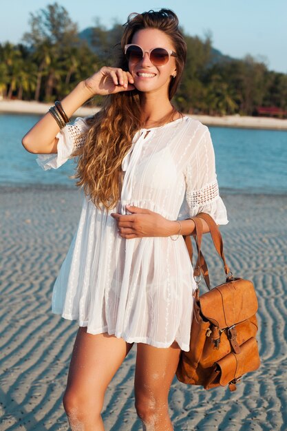 Slim beautiful woman in white dress on tropical beach on sunset holding leather backpack.