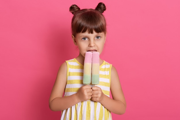 Slim Caucasian girl kid holds two big ice cream looks with her happy eyes, having funny knots, posing isolated over pink wall, female kid biting tasty ice cream.